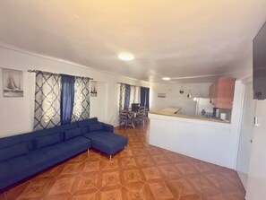 A living room with a blue sectional sofa, a dining area with a table and chairs, and a kitchen area with cabinets and a countertop.