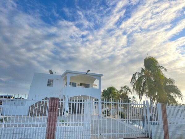 A two-story white building with a satellite dish, a gated entrance, and palm trees in the background.