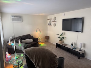 Ein Wohnzimmer mit einer Couch, einem Glastisch, einer Stehlampe, einem Fernseher und einem an der Wand befestigten Regal.