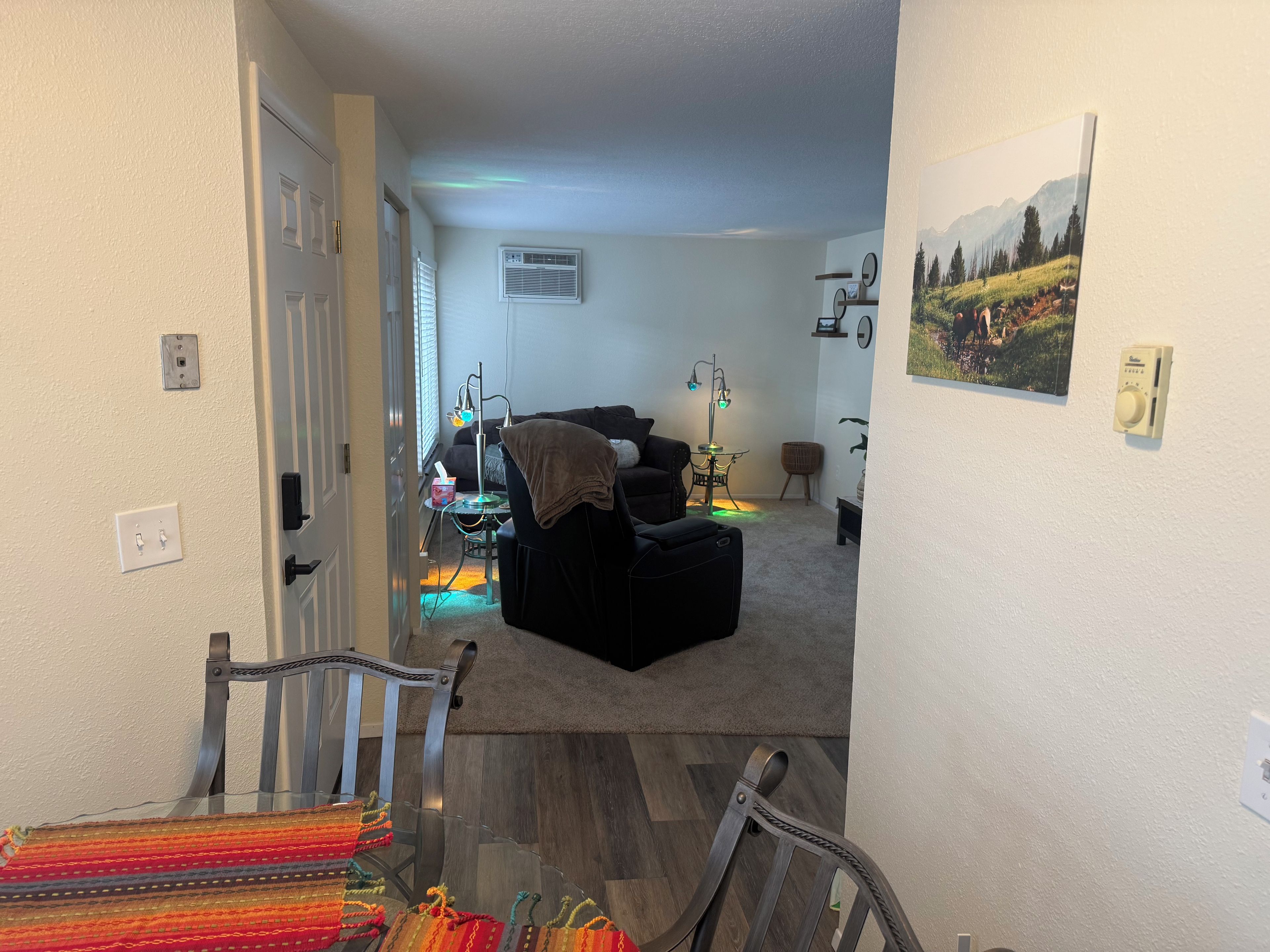 Un salon avec un canapé, un fauteuil, une table et une lampe. Une porte donne accès à une autre pièce.