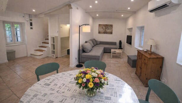 A living room with a grey sofa, a coffee table, and a dining table with a newspaper print tablecloth. There is a staircase leading to another room, and an air conditioning unit on the wall. - Village house for 5 people. (Peyruis)