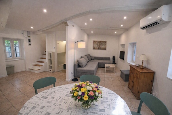 A living room with a grey sofa, a coffee table, and a dining table with a newspaper print tablecloth. There is a staircase leading to another room, and an air conditioning unit on the wall. - Village house for 5 people. (Peyruis)