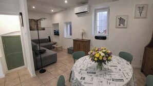 A dining area with a round table covered in a newspaper print tablecloth, surrounded by green chairs. - Village house for 5 people. (Peyruis)
