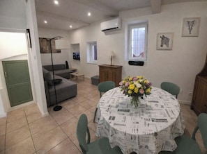A dining area with a round table covered in a newspaper print tablecloth, surrounded by green chairs. - Village house for 5 people. (Peyruis)