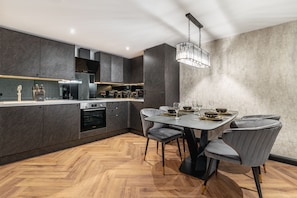 A modern kitchen with a dining area, featuring dark cabinetry, a wooden herringbone floor, and a chandelier.