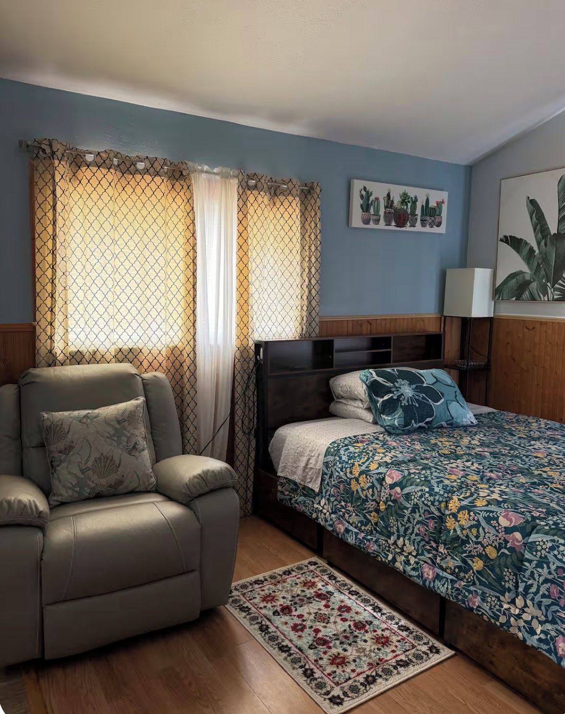 Un dormitorio con cama, una silla, una mesita de noche, una lámpara y una obra de arte en la pared.