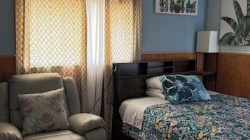 Un dormitorio con cama, una silla, una mesita de noche, una lámpara y una obra de arte en la pared.