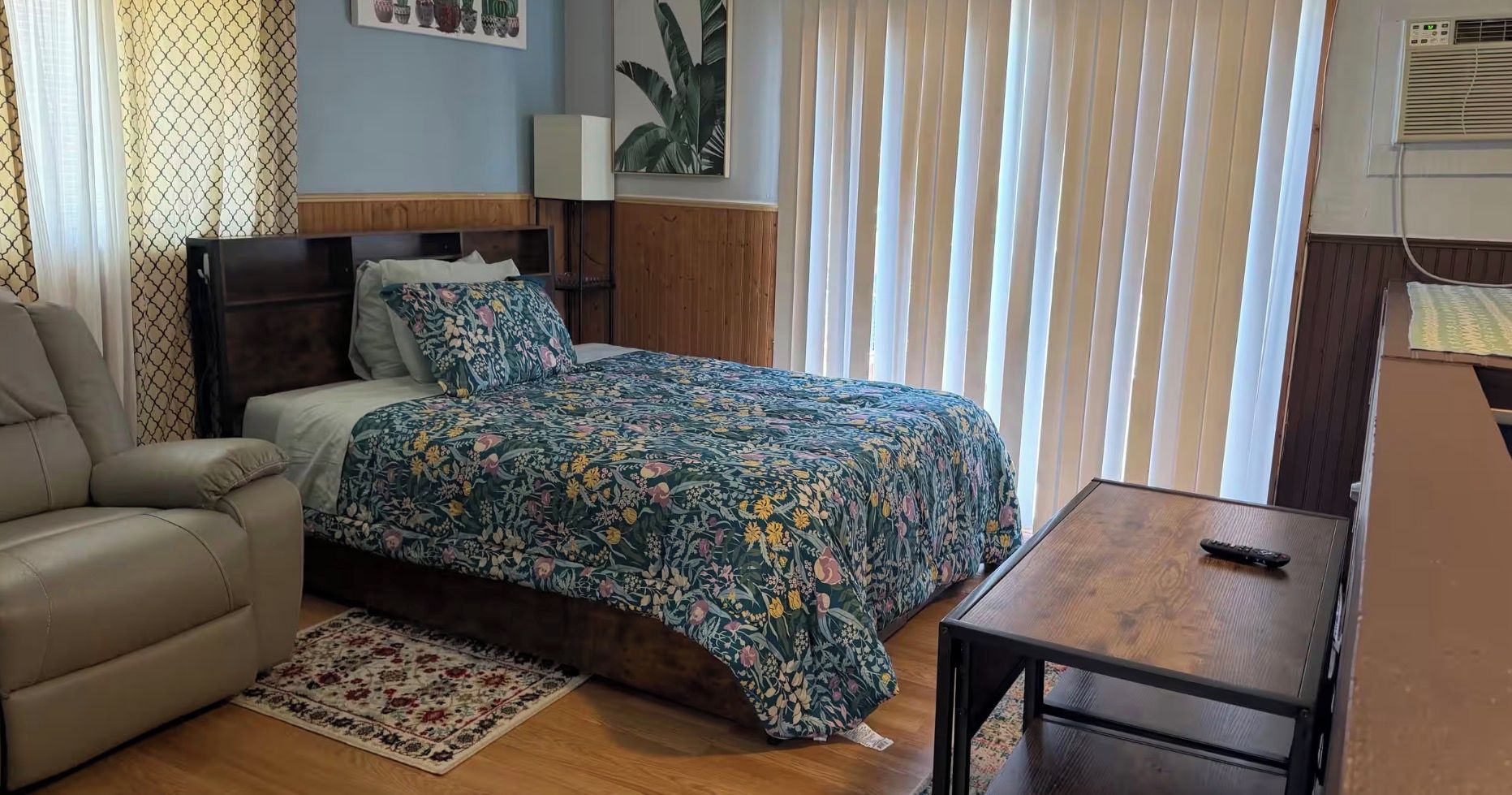 A bedroom with a bed, a sofa, a small table, and a window with blinds.