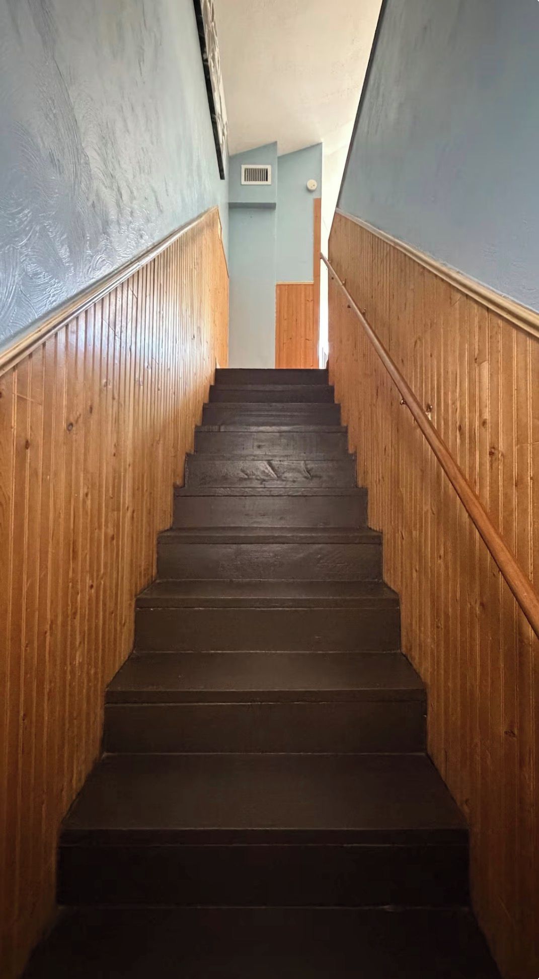 A wooden staircase with a handrail, leading to a landing with a light blue wall and a vent.