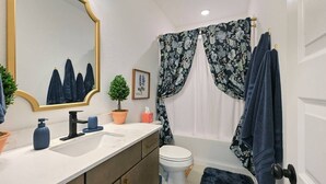 Casa de banho com sanita branca, lavatório com espelho, planta em vaso e cortina de chuveiro com padrão floral.