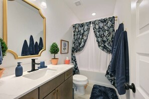 A bathroom with a white toilet, a sink with a mirror, a potted plant, and a shower curtain with a floral pattern. - Pristine New 2BR Townhome | South College Station (College Station)