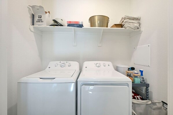 A laundry area with two front-loading washing machines, a shelf with various items, and a whiteboard on the wall.