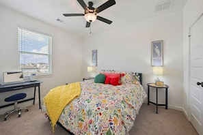 A bedroom with a bed, a desk, a chair, a ceiling fan, and a window with blinds.