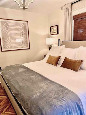 Habitación de hotel con cama, una obra de arte enmarcada, mesita de noche, lámpara y ventana con cortinas.