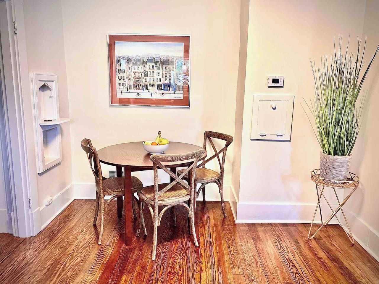 Un coin repas avec une table ronde, quatre chaises, un saladier de fruits, un tableau représentant un paysage urbain encadré, un thermostat fixé au mur et une plante en pot.