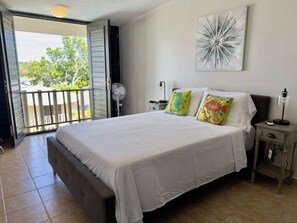 A bedroom with a bed, a nightstand, a wall-mounted lamp, and a window with blinds. - Bright Coastal Stay Near Beach w/ Pool & Tennis (Boquerón)