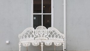 Un banco de metal blanco con diseños intrincados, ubicado frente a una ventana con marco blanco, sobre una pared lisa.