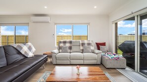 A living room with a sofa, armchair, and ottoman, a coffee table with a vase, and a sliding glass door leading outside. - Woonan - Modern and designed with families in mind (Warrnambool)