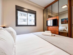 A hotel room with a bed, a window with a view of buildings, a television mounted on a wooden panel, and a painting on the wall. - 1 Bedroom Queen Jardins Pool Garage Metro PKS48 (São Paulo)