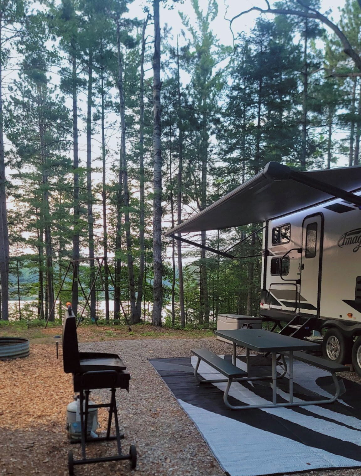 A recreational vehicle (RV) with a covered picnic area, a grill, and a table and chair set up on a gravel surface.