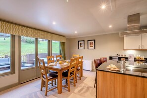A dining area with a table set for four, a kitchen with modern appliances, and a view of a grassy outdoor area through large windows. - The Derwent Spa (Hexham)