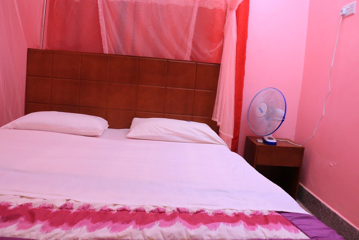 Una cama con ropa blanca, cabecera de madera y mesita de noche con un ventilador.