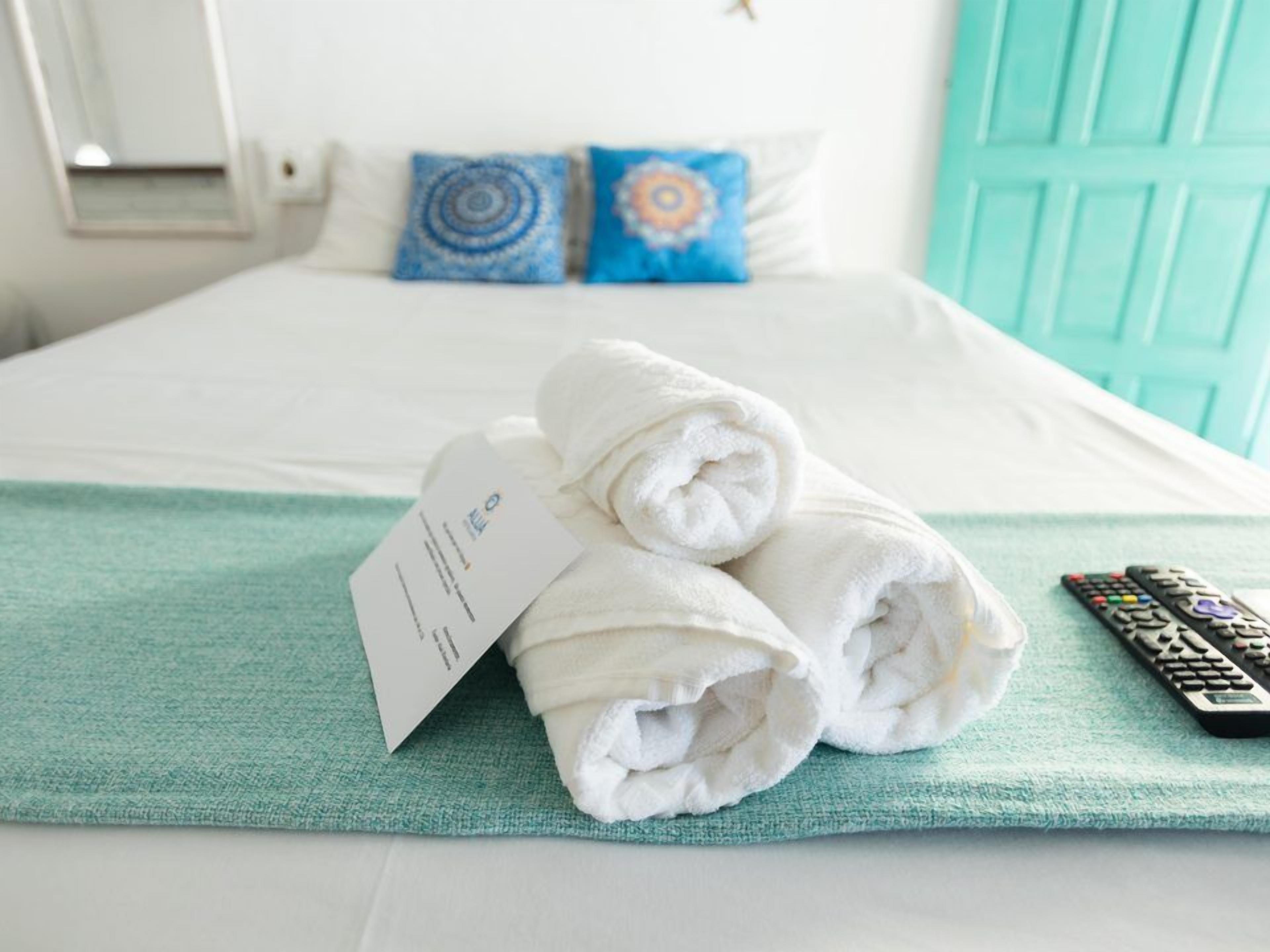 Una cama bien hecha con toallas blancas enrolladas y un control remoto sobre un edredón azul verdoso.