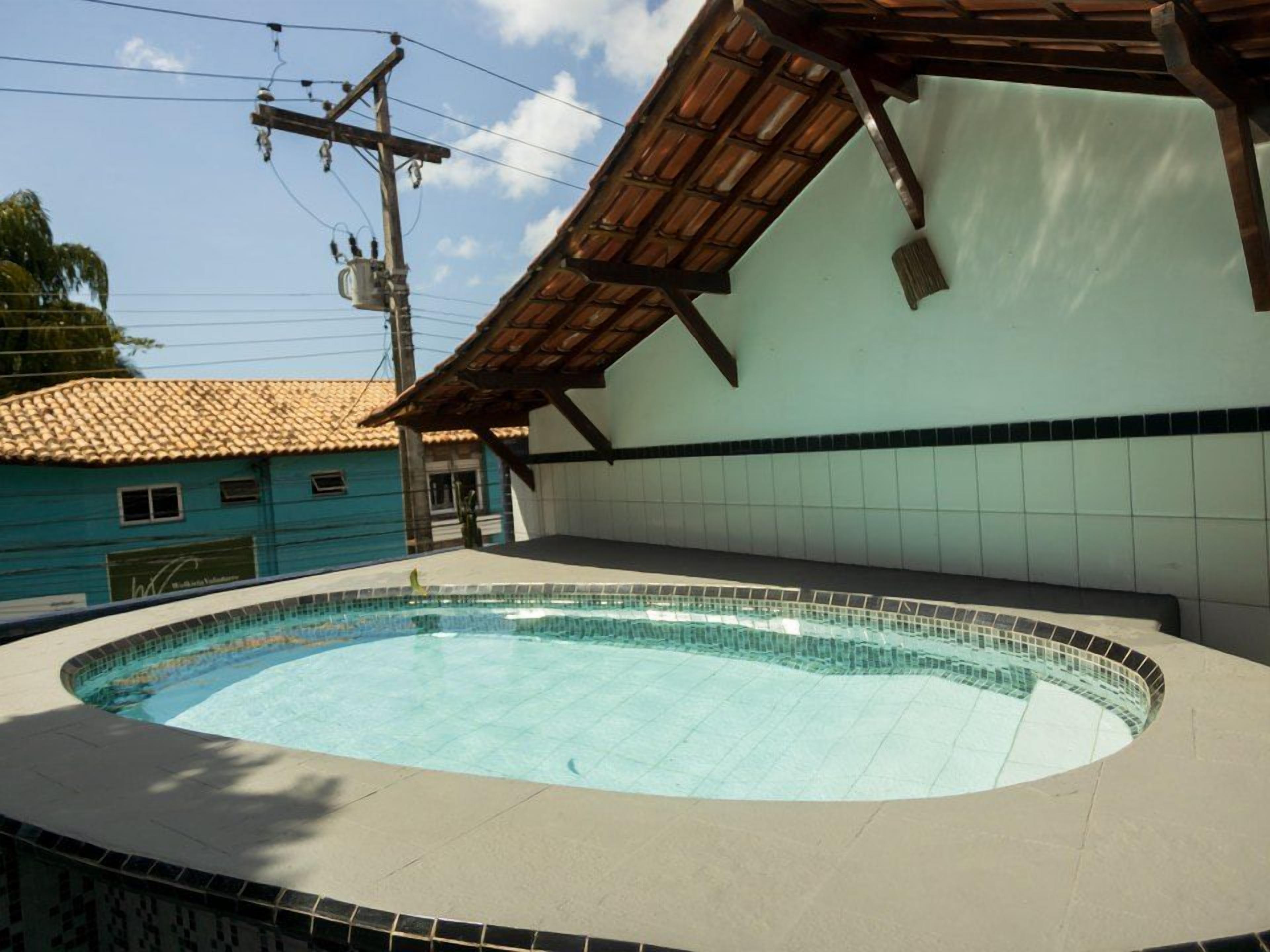 Una piscina con borde de azulejos, un área techada con techo inclinado y un edificio con tejado de azulejos al fondo.