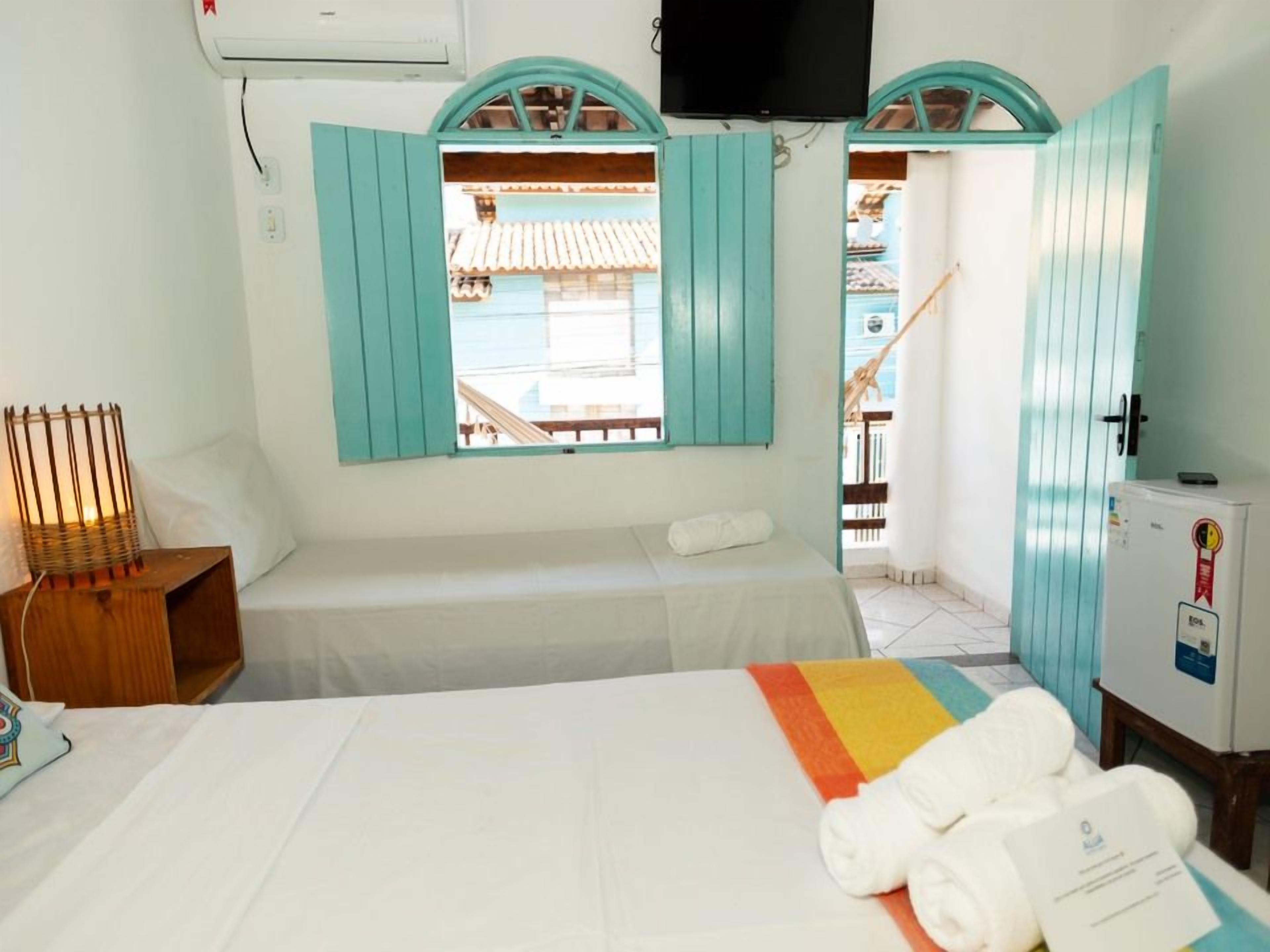 Quarto de hotel com cama, uma mesa pequena, televisão e janela com vista para um edifício.