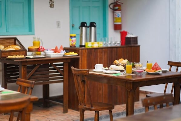 A wooden dining table with breakfast items, a fire extinguisher, and teal-colored doors in the background. - Pousada Coração do Arraial (Porto Seguro)