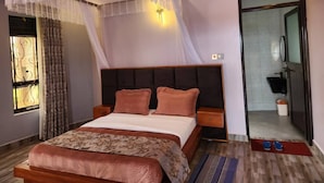 A modern bedroom with a large bed, bedside tables, a canopy, and a view of the bathroom. - Ihunde Farm Lodge (Kiruhura)