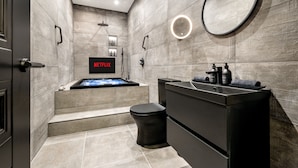 A modern bathroom with a black vanity, a round mirror, a built-in shower, and a wall-mounted Netflix screen.