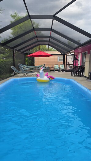Una piscina interior con techo transparente, rodeada de sillones de descanso y un paraguas rojo.
