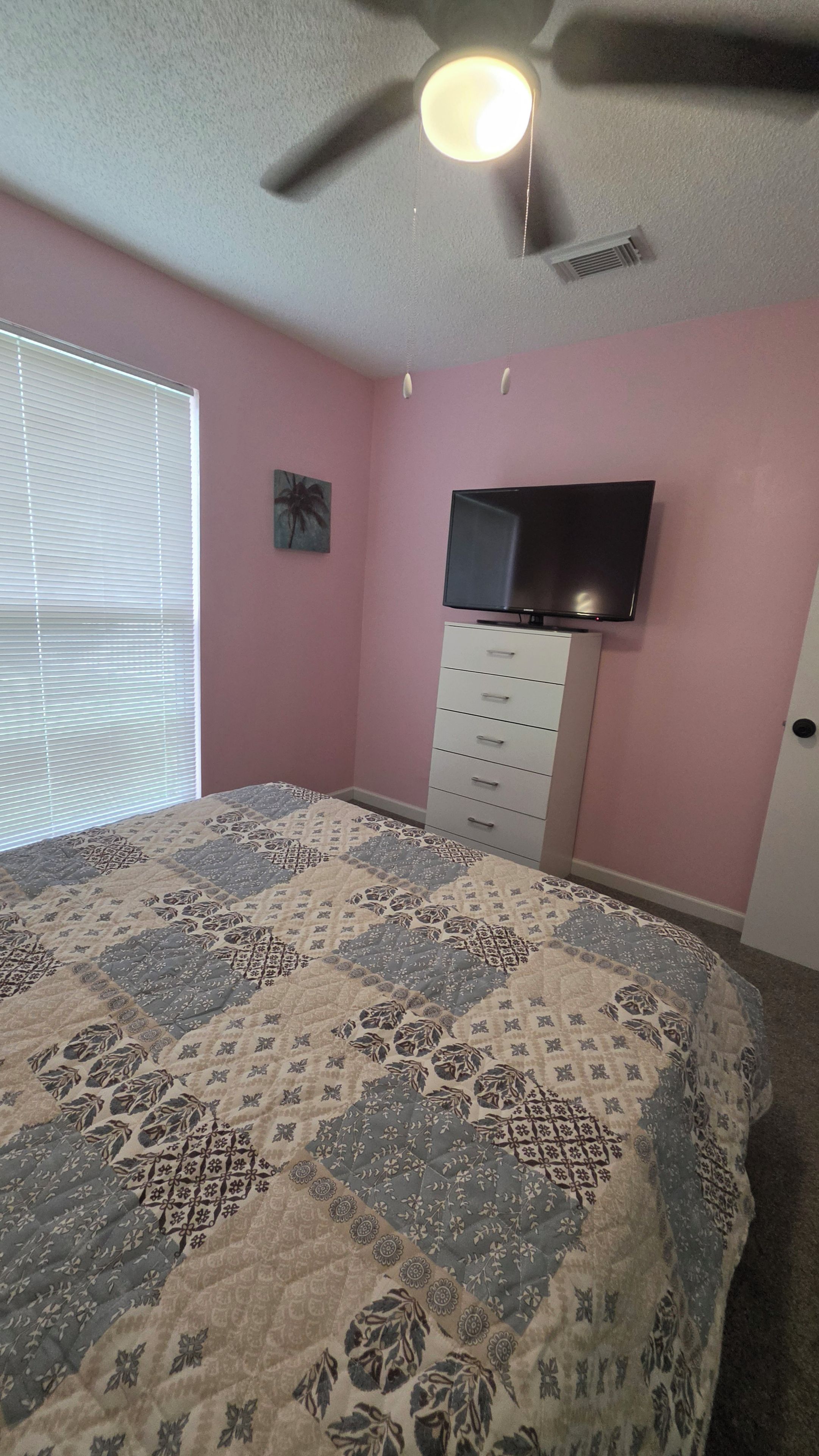 Une chambre à coucher avec un lit, un ventilateur de plafond, un téléviseur fixé sur une commode et une fenêtre avec des stores.