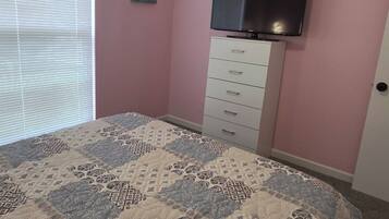 Une chambre à coucher avec un lit, un ventilateur de plafond, un téléviseur fixé sur une commode et une fenêtre avec des stores.
