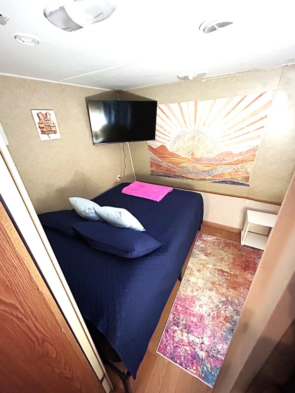 A small room with a bed, a flat-screen TV mounted on the wall, a colorful tapestry, and a small white table.