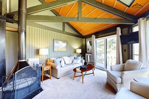 A cozy living room with a sofa, a coffee table, and a fireplace. There is a window with curtains and a view of the outdoors. - Sleep at The Slopes! 2 Bedroom Round House Slopesider #1 (Sugar Mountain)