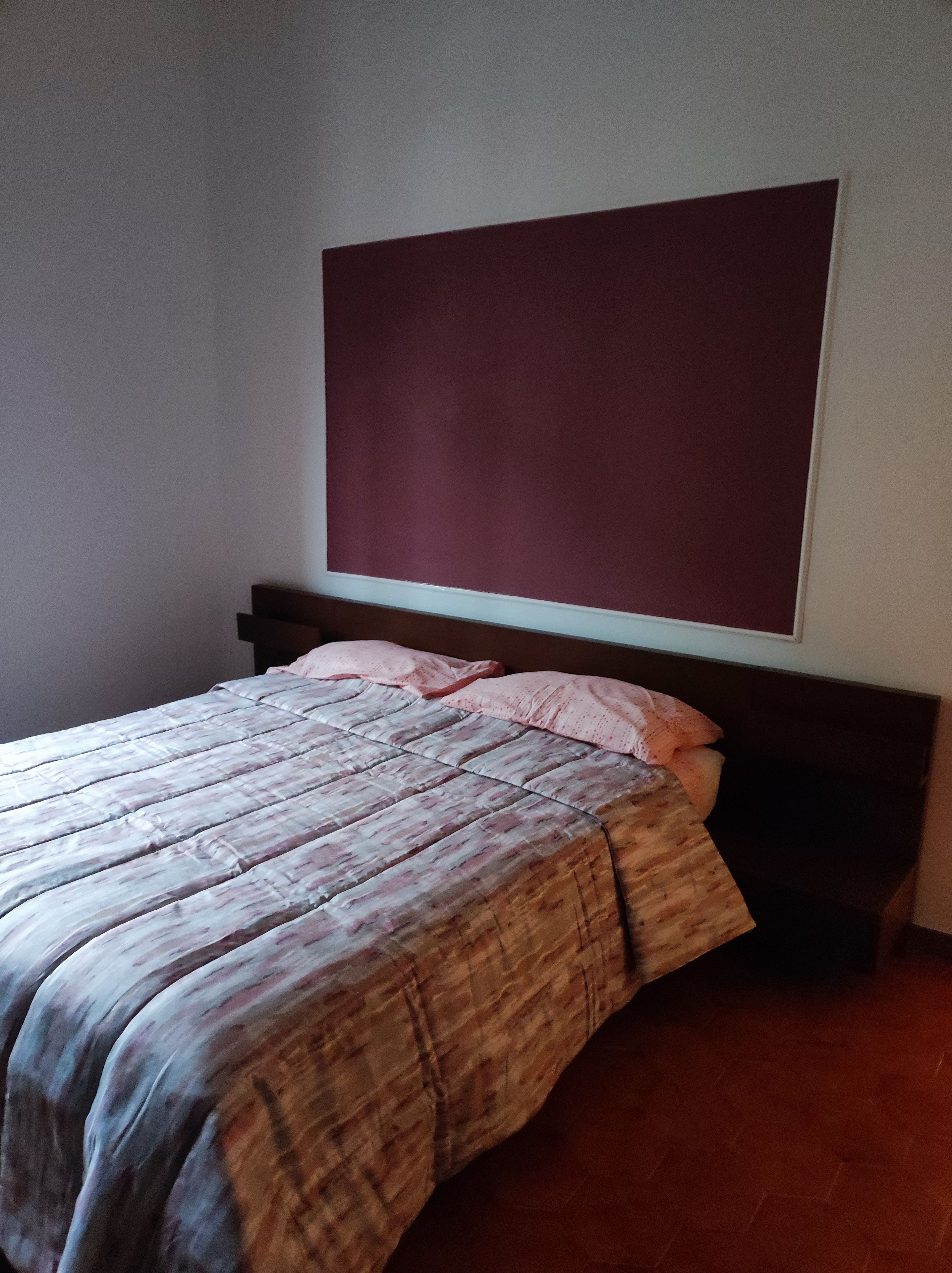 A bed with a patterned bedspread and pillows, a dark headboard, and a large, plain wall above.