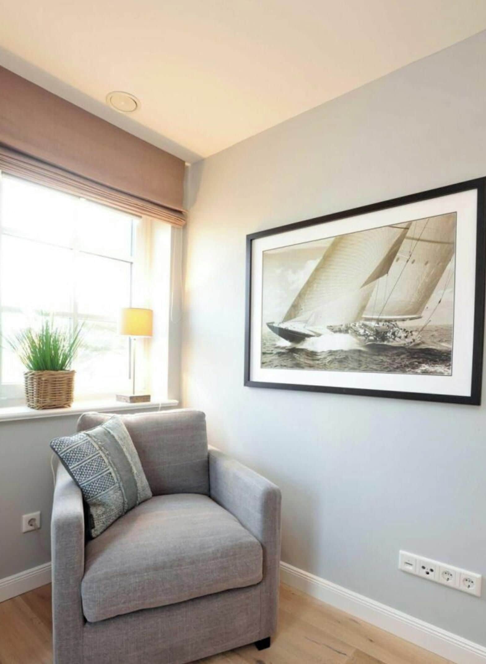 Una habitación con un sillón gris, un cuadro enmarcado de un velero, una ventana con una planta y una lámpara.