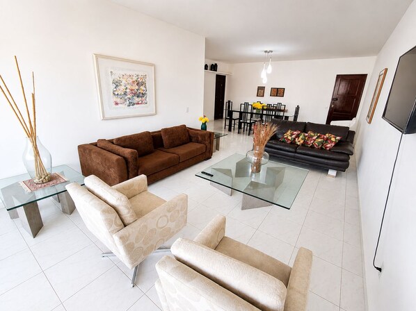 Ein modernes Wohnzimmer mit einem braunen Sofa, beigefarbenen Sesseln und einem Glastisch. Im Hintergrund befindet sich ein Essbereich mit einem schwarzen Sofa und einem Esstisch.