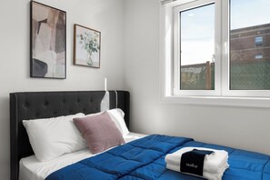 Ein Schlafzimmer mit einem Bett, Kissen, einer blauen Bettdecke, einem Fenster mit Blick und zwei gerahmten Kunstwerken an der Wand.
