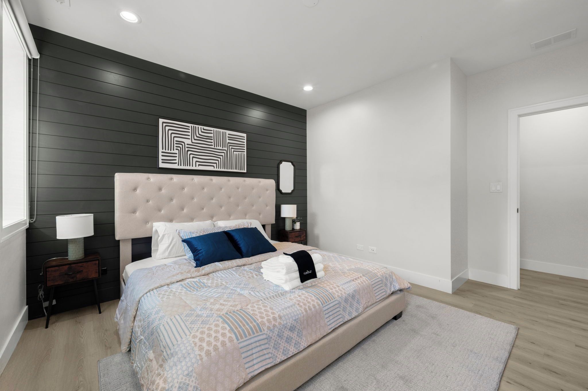 A modern bedroom with a tufted headboard, a large bed with patterned bedding, a bedside table with a lamp, and a wall-mounted artwork.