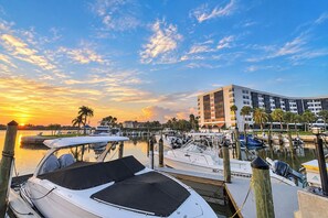 Un port de plaisance avec des bateaux amarrés, un bâtiment moderne à plusieurs étages, et un coucher de soleil en arrière-plan.