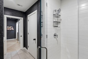 Uma casa de banho moderna com azulejos brancos tipo metrô, cabine de duche em vidro, porta preta e um chuveiro com um cesto de casa de banho prateado.