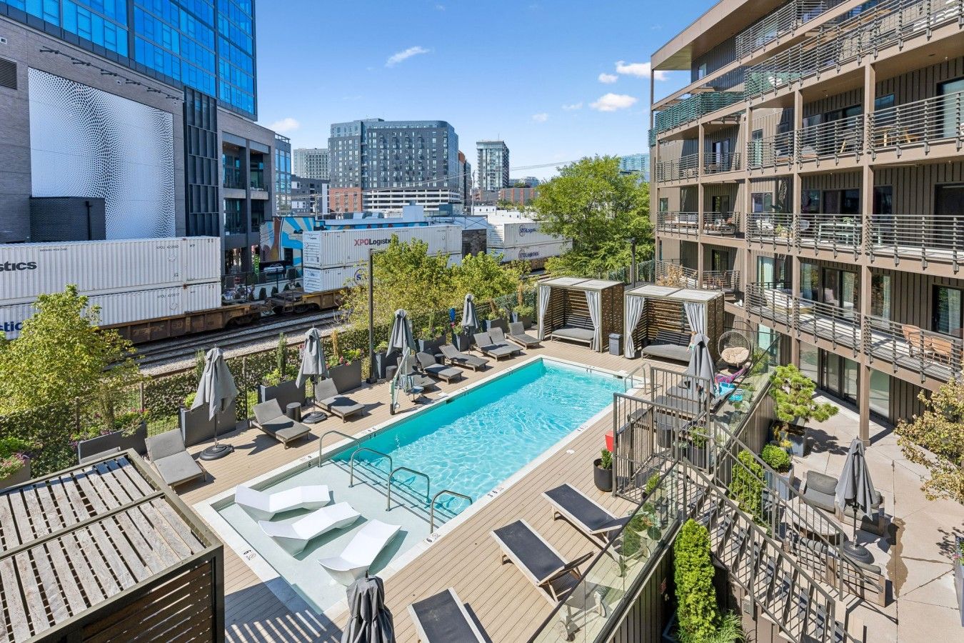 A rooftop pool area with lounge chairs and umbrellas, surrounded by modern buildings and a clear sky.