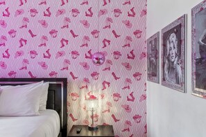 Quarto com papel de parede estampado, cama com roupa de cama branca, mesa de cabeceira com abajur e quadros nas paredes.
