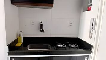 Uma cozinha compacta com bancada preta, paredes com azulejos brancos, armário preto, pia, fogão e lixeira.