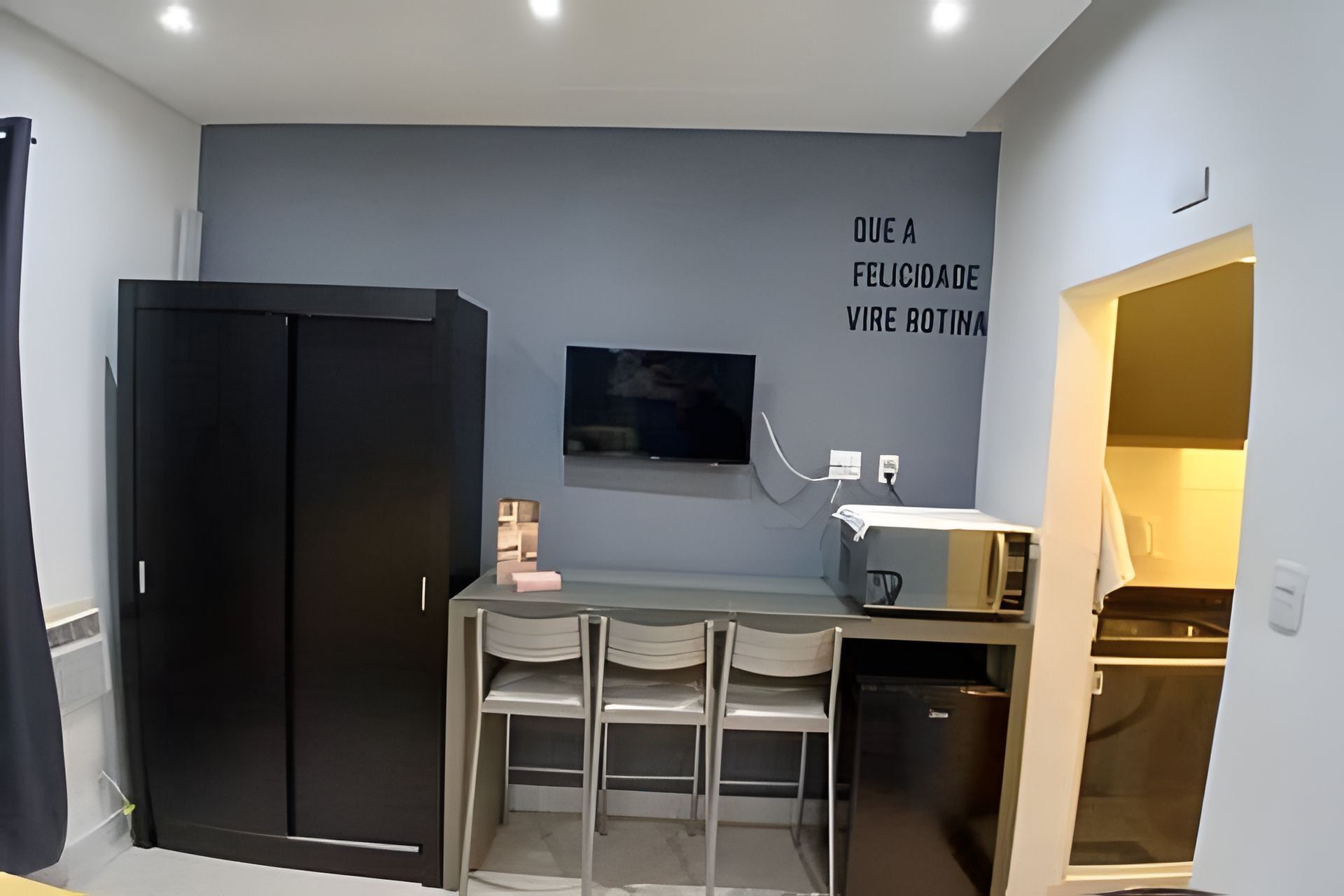 Una cocina compacta con un refrigerador negro, un pequeño comedor con sillas, un microondas y un televisor montado en la pared.