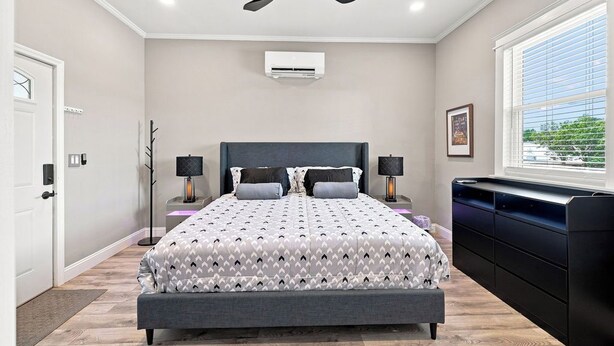 Une chambre à coucher comprenant un lit, des tables de chevet, un ventilateur de plafond, un appareil de climatisation, une fenêtre avec des stores et une vue sur la verdure à l’extérieur.