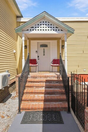 A two-story building with a porch, a door marked '102', and two red chairs.